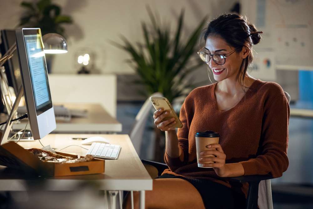 femme d'affaires effectuant un appel vidéo avec son téléphone portable tout en travaillant sur son ordinateur, assise dans un bureau.